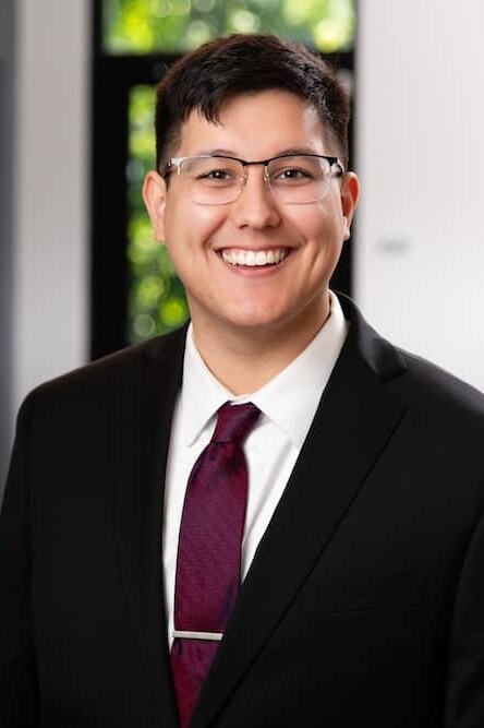 Smiling man with glasses and short, dark hair wearing a suit with dark red tie