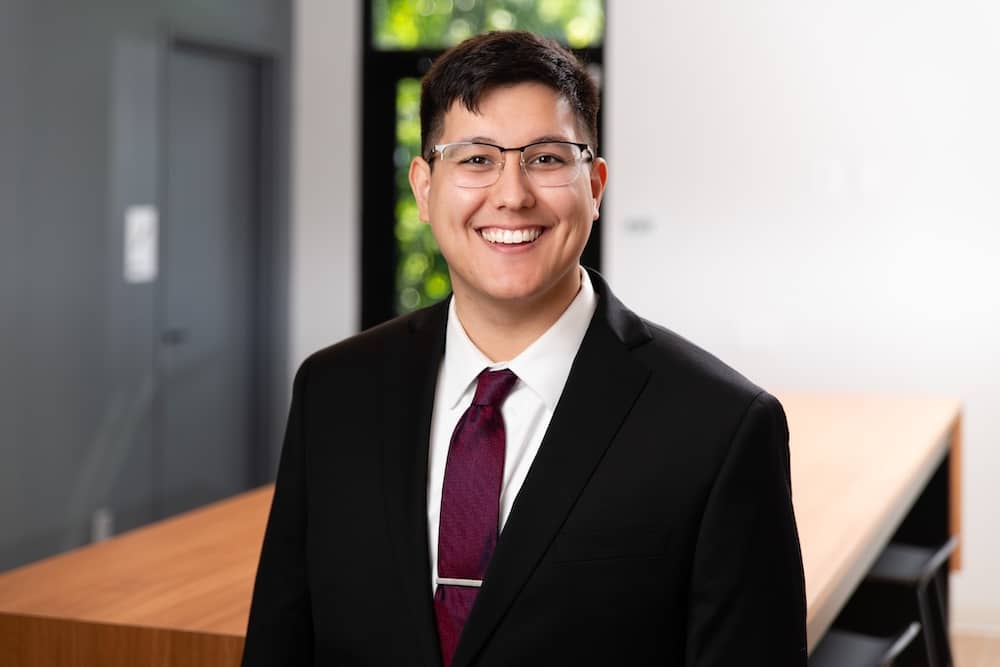 Smiling man with glasses and short, dark hair wearing a suit with dark red tie